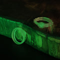 Lifestyle photo of two translucent glow-in-the-dark Slay the Spire Snake Rings on dark wood block emitting vivid green glow in low light