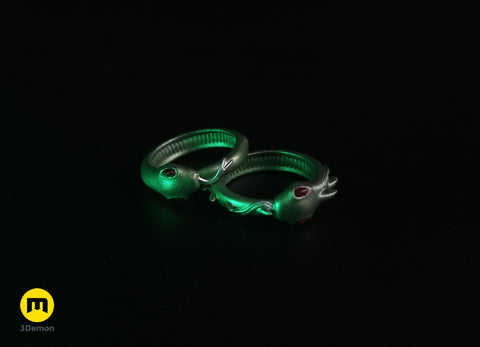 Lifestyle photo of two 3D printed Slay the Spire Snake Rings in metallic green on black surface showing snake head with red eyes silver body detail