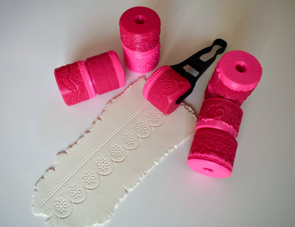 Group flat lay of multiple pink 3D printed lace texture wheels with black handle where one pink daisy lace pattern wheel actively rolls embossed daisy design into white clay
