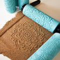Top-down photo showing three distinct large mandala imprints in brown clay slab with two turquoise 3D printed texture rollers placed beside results