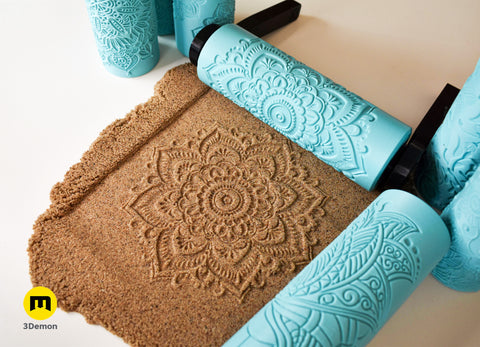 Top-down photo showing three distinct large mandala imprints in brown clay slab with two turquoise 3D printed texture rollers placed beside results