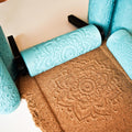 3D printed turquoise mandala texture roller with black handle actively imprinting detailed floral mandala pattern into flat slab of brown kinetic sand