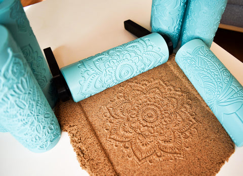 3D printed turquoise mandala texture roller with black handle actively imprinting detailed floral mandala pattern into flat slab of brown kinetic sand