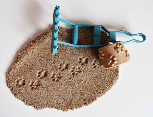 Lifestyle photo of child hand holding teal 3D printed multi-tool handle with beige texture roller attached rolling animal paw print patterns into sandbox sand from diagonal angle above