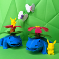 Toy frogs with flower structures and butterflies on a green geometric-patterned background with pikachu in front