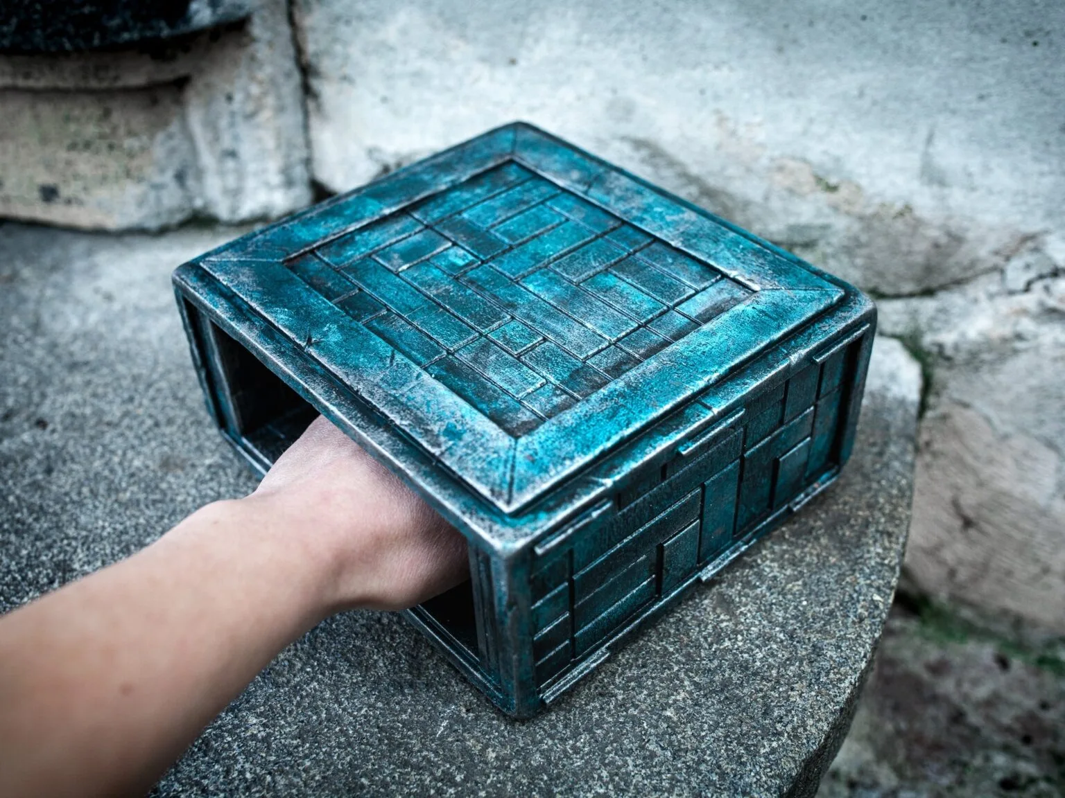 Hand photo of Box of Pain 3D printed model, turquoise textured metal finish, held outdoors