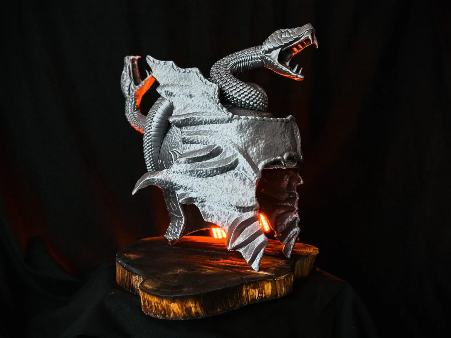 Lifestyle photo of a grey 3D printed Messmer the Impaler helmet with snake details on a wooden base.