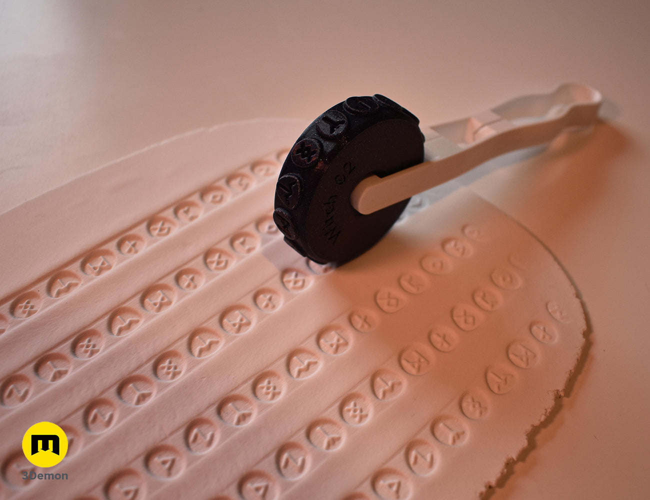 Black 3D printed stamp wheel with a white handle creating rows of skull patterns on white clay.