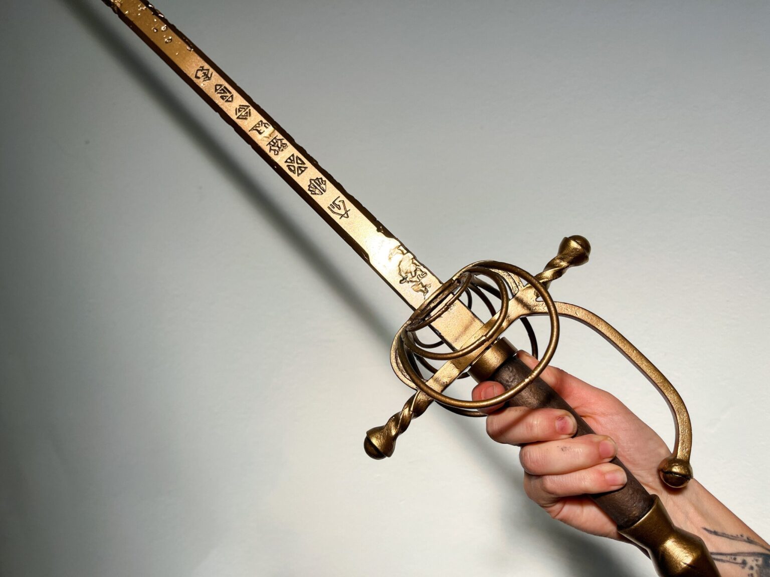 Lifestyle close-up photo of hand holding painted Expedition 33 Maellum rapier hilt in workshop setting showing golden cage guard and cream leather-wrapped grip with metallic sheen